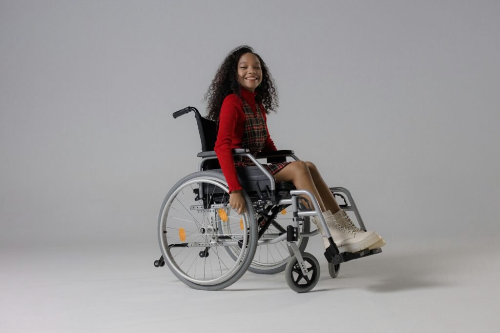 Mobility Aid Training with Kids Young girl smiling in a wheelchair, wearing a plaid dress and boots against a white background.