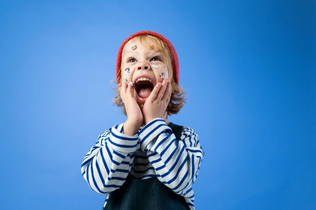 pexels-photo-4715322-4715322 Happy child with face stickers and red hat on a blue background, expressing joy and surprise.